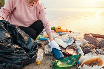 Young woman collects plastic garbage in trash bag on the sandy beach of sea at sunset. Spilled garbage on beach. Empty used dirty plastic bottles. Environmental pollution the Black Sea coast