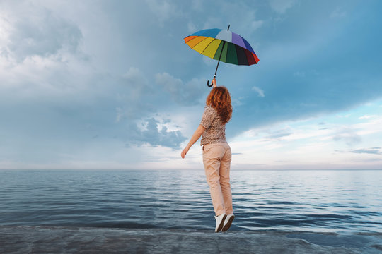 Silhouette of a young woman jumping up from happiness on the embankment in the seaport with a multicolored umbrella against the backdrop of sunset and rainbow, with a cloudy rainy sky and blue sea