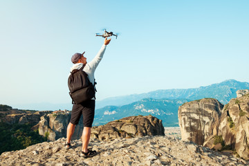 Obraz premium Young man photographer stands on top of mountain in Meteora, Greece and catches drone in the hand with which he shot panorama of mountains and monasteries. Summer tourism. Copy space