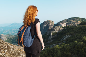 Relaxed red-haired woman tourist backpack stands on top of high mountain against the backdrop of the Meteora Mountains in Greece, and looks out over the mountains on the horizon in summer. Copy space