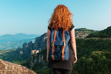 A relaxed red-haired woman with a tourist backpack stands on top of a high mountain against the backdrop of the Meteora Mountains in Greece, and looks out over the mountains on the horizon in summer