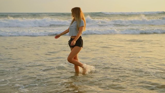 Young female felling fun and free and walking barefoot on beach at sunset. Carefree girl on tropical beach runs into refreshing sea at golden sunrise. Happy tourist woman on summer vacation.
