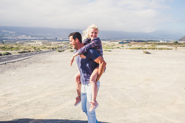 Happiness and outdoor funny leisure activity for young laughig people together - man carrying blonde beautiful woman with road and desert in background