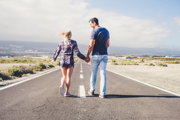 Fototapeta premium life together concept with two young people viewed from back walking together on a long straight road taking holding hands and smiling - focus on asphalt in foreground