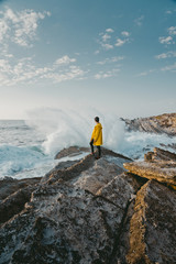 Traveler in the yellow jacket standing on the rocks in front of a huge waves