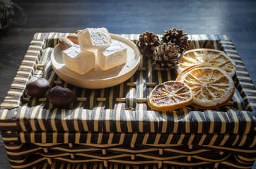 Delicious white Turkish delight on a wooden plate sprinkled with powdered sugar. Oriental pieces of sweets next to cones, chestnuts and dried orange rings. Winter christmas dessert in the sunshine