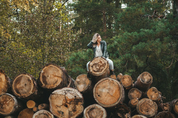Beautiful woman in leather jacket sitting on stack of large logs in autumn park