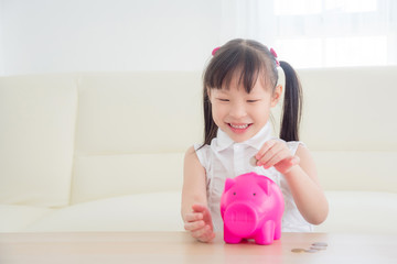Little asian girl inserting coin into pink piggy bank and smiles.