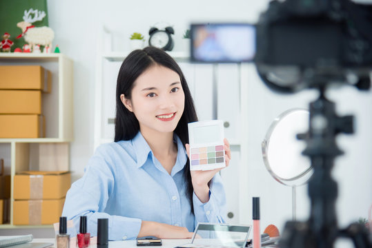 Beautiful asian girl holding eyeshadow palette talking about her daily make-up routine in front of camera to recording vlog video live streaming at her shop.