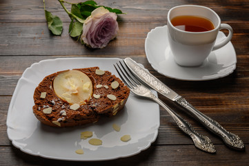 Piece of homemade chocolate pear cupcake with ginger and cardamom on a white plate. Knife and fork and tea in a white cup and saucer. The rose bud in the background