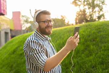 Naklejka na ściany i meble Portrait of a positive young man with a mustache and beard listening to an audiobook while walking through the park. Leisure learning concept.