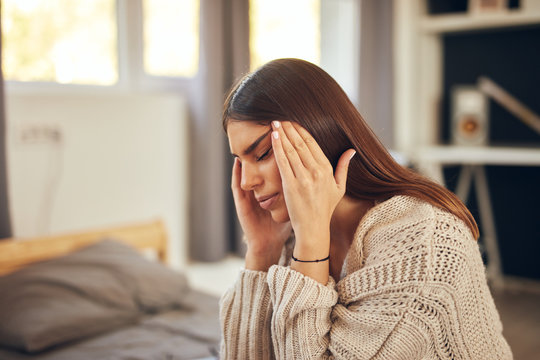 Attractive caucasian brunette dressed in beige sweater sitting on bed in bedroom and having headache.