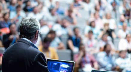 Speaker giving a talk on corporate business conference. Unrecognizable people in audience at...
