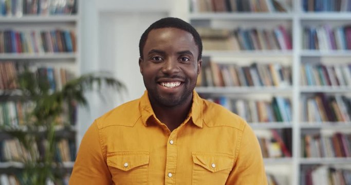 Portrait of the African American attractive young man in yellow shirt smiling happily to the camera in the library. Close up.