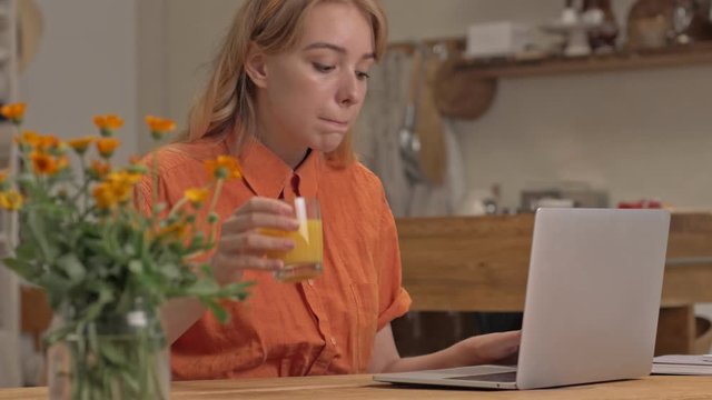 Concentrated young blonde woman working on laptop and drinking juice while sitting at the kitchen
