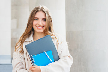 Young blonde girl student smiling against university. Cute girl student holds folders and notebooks...