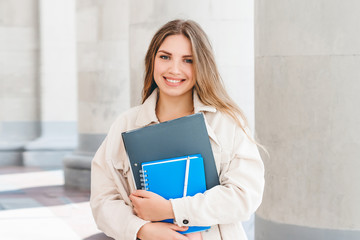Young blonde girl student smiling against university. Cute girl student holds folders and notebooks...