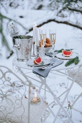 Decorated table for a romantic dinner with candles, sparkling wine and fruit in the winter forest