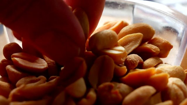Human hand takes roasted and salted peanuts from a small transparent bowl