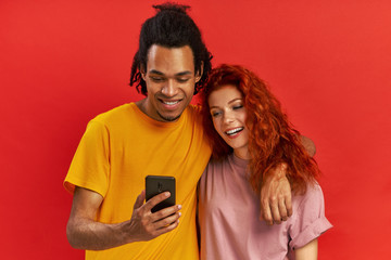 Positive young mixed race couple embrace each other, look joyfully at mobile phone, glad to view common photos, satisfied with good wireless connection, against red background. Relationship concept.