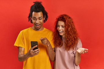 Optimistic young ginger female and Afroman users of smart technology, feel good from successful cellphone updating, gaze at screen, check article, wear casual clothes, stand over red background.