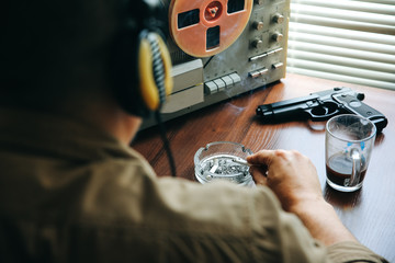 Special agent listens on the reel tape recorder. Officer is smoking a cigarette. KGB spying on conversations. Hand with cigarette near the ashtray. Gun on the table.