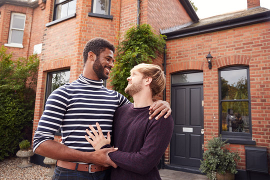 Excited Gay Male Couple Standing Outside New Home On Moving Day Together