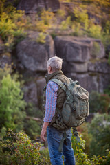 Back view of tourist man with a gray beard with a backpack on his shoulders against the backdrop of the gorge, rocks and stones, the concept of tourism and outdoor activities in old age