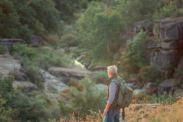 Fototapeta Back view of tourist man with a gray beard with a backpack on his shoulders against the backdrop of the gorge, rocks and stones, the concept of tourism and outdoor activities in old age obraz