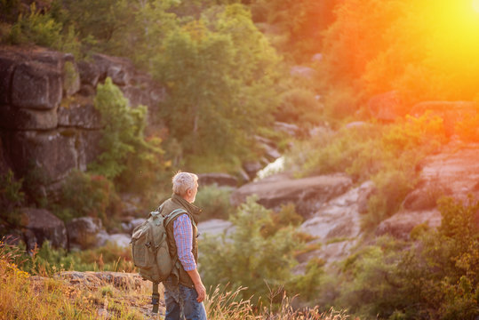 Back view of tourist man with a gray beard with a backpack on his shoulders against the backdrop of the gorge, rocks and stones, the concept of tourism and outdoor activities in old age
