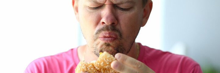 Portrait of unsatisfied male eating tasteless hamburger with obvious disgust. Bearded person holding hideous and sickening burger in hands. Blurred background