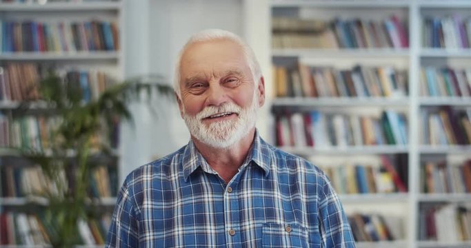 Portrait of the senior Caucasian man with grey hair and in the blue motley shirt smiling cheerfully in the library. Close up.