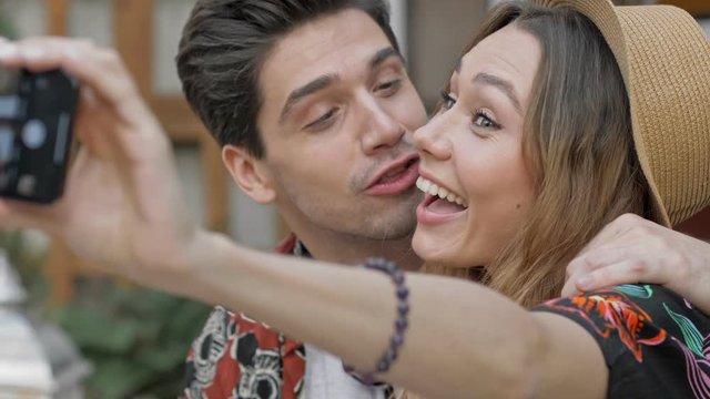 Close up view of smiling young lovely couple winking and gesturing with hands while taking selfie photos on smartphone
