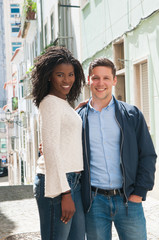 Sweet couple enjoying weekend outside. Young man and woman standing for camera in old town street, hugging and smiling. Diverse couple concept