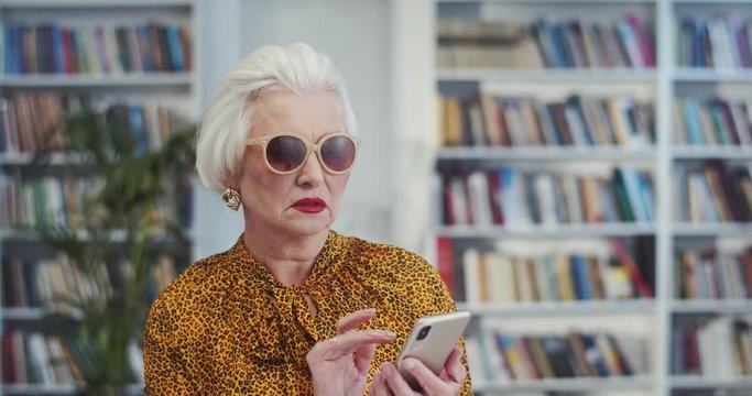Old fancy and beautiful Caucasian woman in sunglasses and with grey hair texting a message on the smartphone in the library. Close up.