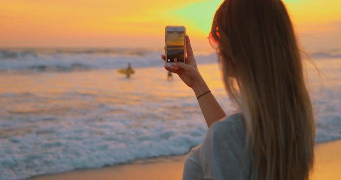 Young female felling fun and free on beach at sunset. Carefree girl on tropical beach takes photos of nature on a smartphone at golden sunrise. Happy tourist woman on summer vacation. 5g Internet.