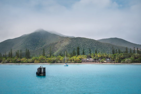 View of Pic N'Ga Mountain engulfed in clouds and the turquoise waters of Kuto Bay with Pine and Coconut palm trees and a waterfront restaurant on its white beach on Isle of Pines in New Caledonia