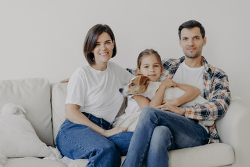 Friendly family pose together at sofa, enjoys domestic atmosphere. Father, mother, their little daughter and pedigree dog spend weekend at home, pose in living room, have happy face expressions