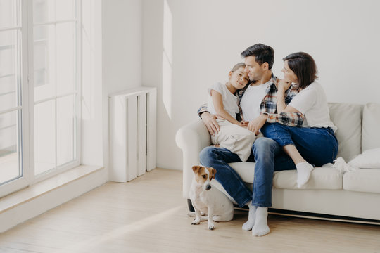 Indoor shot of happy mother and father spend weekend with small daughter, pose all together on comfortable sofa, have fun and pleasant talk with each other. Little dog sits near on floor. Family