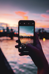 View through a smart phone screen with blurry background of a city at colorful sunset light. A girl...