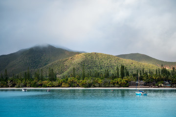 Kuto Bay View with turquoise waters, a white sandy beach with a waterfront restaurant and Pic N'Ga Mountain top engulfed in clouds and sun-lit mountain ridges on Isle of Pines, New Caledonia.