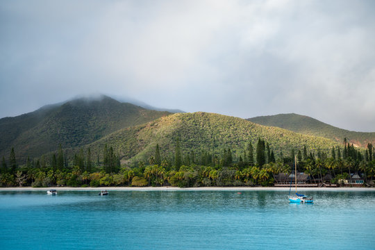 Kuto Bay View with turquoise waters, a white sandy beach with a waterfront restaurant and Pic N'Ga Mountain top engulfed in clouds and sun-lit mountain ridges on Isle of Pines, New Caledonia.