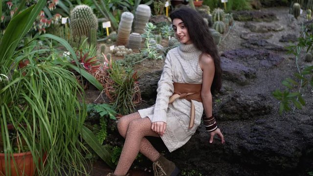 Attractive smiling lady with long dark hair is posing on big stone in Botanical garden