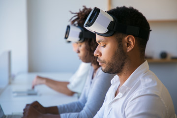 Team of VR developers working on project. Man and women wearing VR headset, sitting at long desk,...