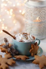 Hot winter drink: chocolate with whipped cream in blue mug. Christmas time. Cozy home atmosphere, white background. Homemade gingerbread cookies, cones, candle, lights as decor. Holiday festive mood