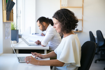 Obraz premium Satisfied female professional working in office. Middle aged Caucasian woman sitting at workplace, using laptop, looking at monitor, smiling. Working in office concept
