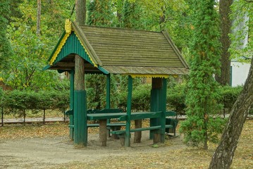 gray wooden gazebo with a green table and chairs among the vegetation and trees in the park