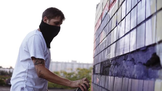 Graffiti artist painting with aerosol. Man in balaclava and white T shirt with spray bottle. Slow Motion. Young urban painter with covered face drawing colorful graffiti on the urban street wall at
