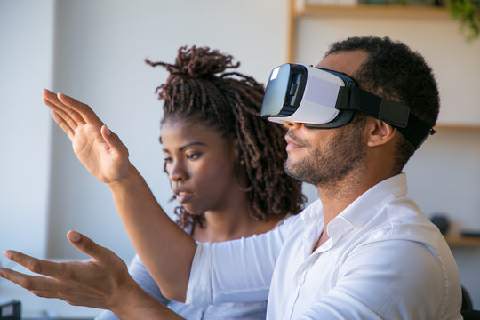 Diverse colleagues testing VR simulator together. Young woman sitting at male colleague wearing virtual reality glasses, touching air. VR technology concept