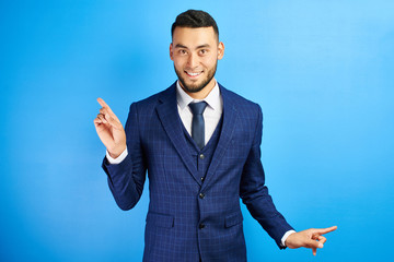 Asian Kazakh man in a business suit smiles friendly and shows fingers to the sides on copy space on a blue wall isolated